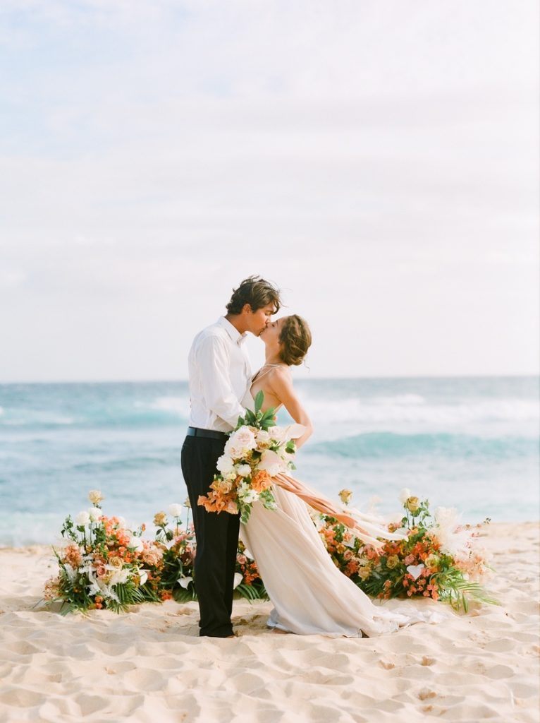 A couple kissing at a beach wedding surrounded by tropical flowers